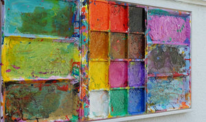 Das Gemälde "Unterwegs" ist ein überdimensionierter, benutzter Farbkasten 130 x 60 x 5 cm groß. Das Kunstwerk ist mit einem weißen Rahmen versehen (ca 2 cm Breite). In den Mischfächer sind Blau, Violett, Grün, Braun und Gelb zu sehen. In der Mitte befinden sich 12 Farbfächer mit den üblichen Farben einer Palette. In den Zwischenräumen sind Farbkleckse und -flecken sichtbar. Modernes Bild als Wandskulptur. ARTLET | Atelier Hellbusch