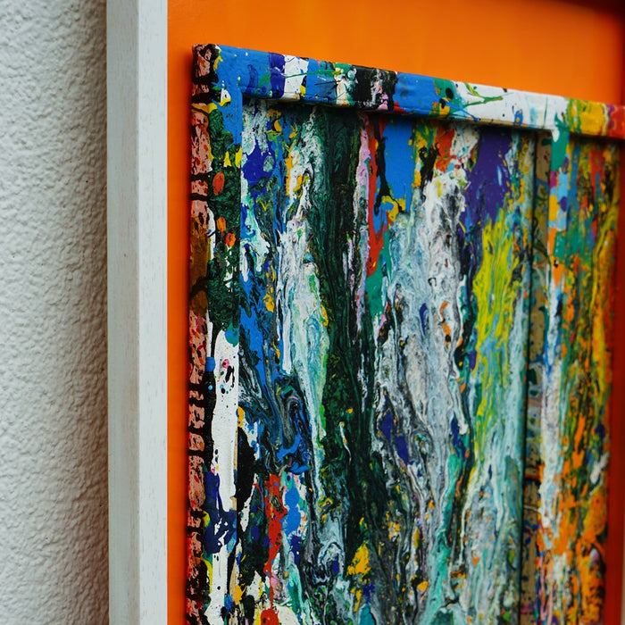 Quadratisches Kunstwerk RE 20 mit intensiven Farbschlieren in Grün, Blau, Orange und Weiß, eingefasst in weißem Rahmen mit leuchtend orangem Hintergrund – Magie aus den Resten eines Farbexperiments (Seitenaufnahme). Atelier Hellbusch.