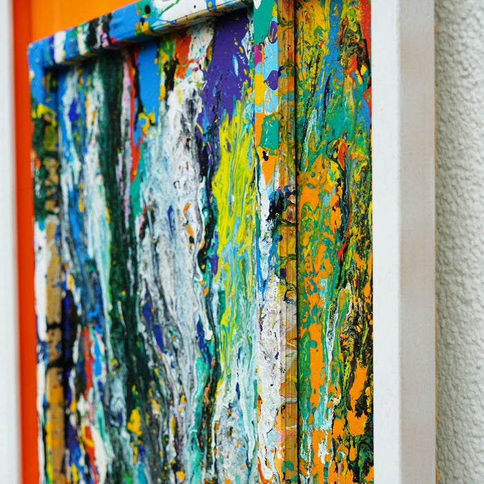 Quadratisches Kunstwerk RE 20 mit intensiven Farbschlieren in Grün, Blau, Orange und Weiß, eingefasst in weißem Rahmen mit leuchtend orangem Hintergrund – Magie aus den Resten eines Farbexperiments (Seitenaufnahme). Atelier Hellbusch.