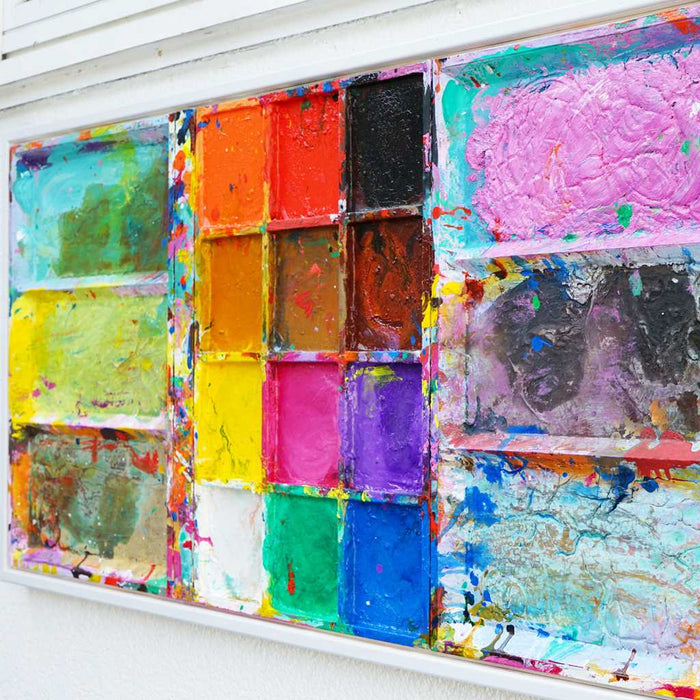 Das Gemälde "Unterwegs" (Seitenansicht) ist ein überdimensionierter, benutzter Farbkasten 130 x 60 x 5 cm groß. Das Kunstwerk ist mit einem weißen Rahmen versehen (Schattenfuge, weiß, ca 2 cm Breite). In den Mischfächer sind Blau, Violett, Grün, Braun und Gelb zu sehen. In der Mitte befinden sich 12 Farbfächer mit den üblichen Farben einer Palette. In den Zwischenräumen sind Farbkleckse und -flecken sichtbar. Modernes Bild als Wandskulptur. ARTLET - Atelier Hellbusch