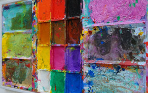 Das Gemälde "Unterwegs" ist ein überdimensionierter, benutzter Farbkasten 130 x 60 x 5 cm groß. Das Kunstwerk ist mit einem weißen Rahmen versehen (ca 2 cm Breite). In den Mischfächer sind Blau, Violett, Grün, Braun und Gelb zu sehen. In der Mitte befinden sich 12 Farbfächer mit den üblichen Farben einer Palette. In den Zwischenräumen sind Farbkleckse und -flecken sichtbar. Modernes Bild als Wandskulptur. ARTLET | Atelier Hellbusch